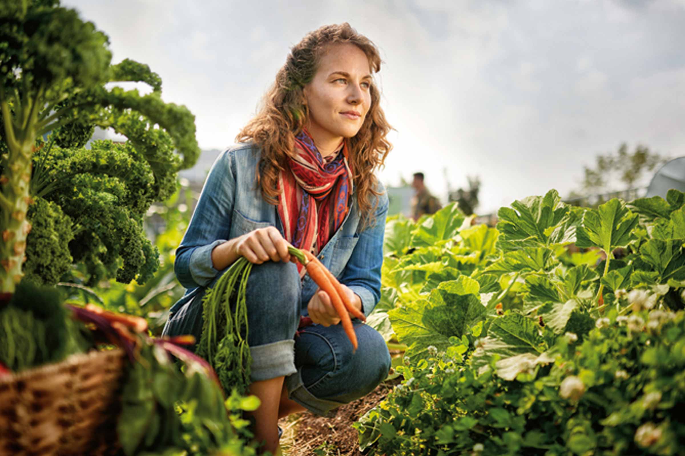 Een vrouw zit gehurkt in een tuin en houdt vers geoogste wortelen vast.