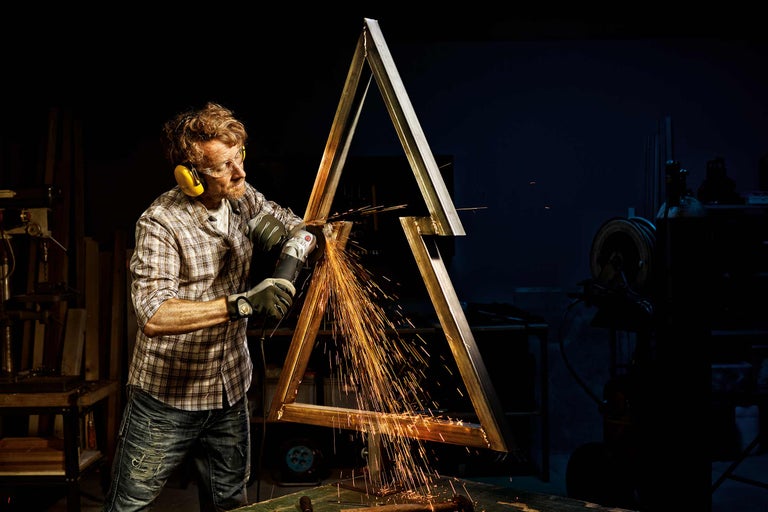 Man werkt met een haakse slijper aan een metalen frame in de vorm van een driehoek, vonken vliegen eraf.