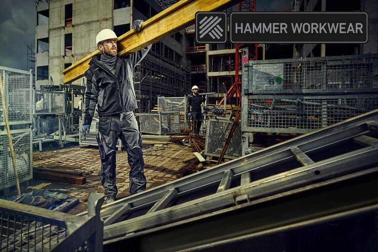 Een bouwvakker draagt een houten balk op een bouwplaats. Op de achtergrond staat een onafgewerkt gebouw. Afgebeeld is Hammer Workwear.