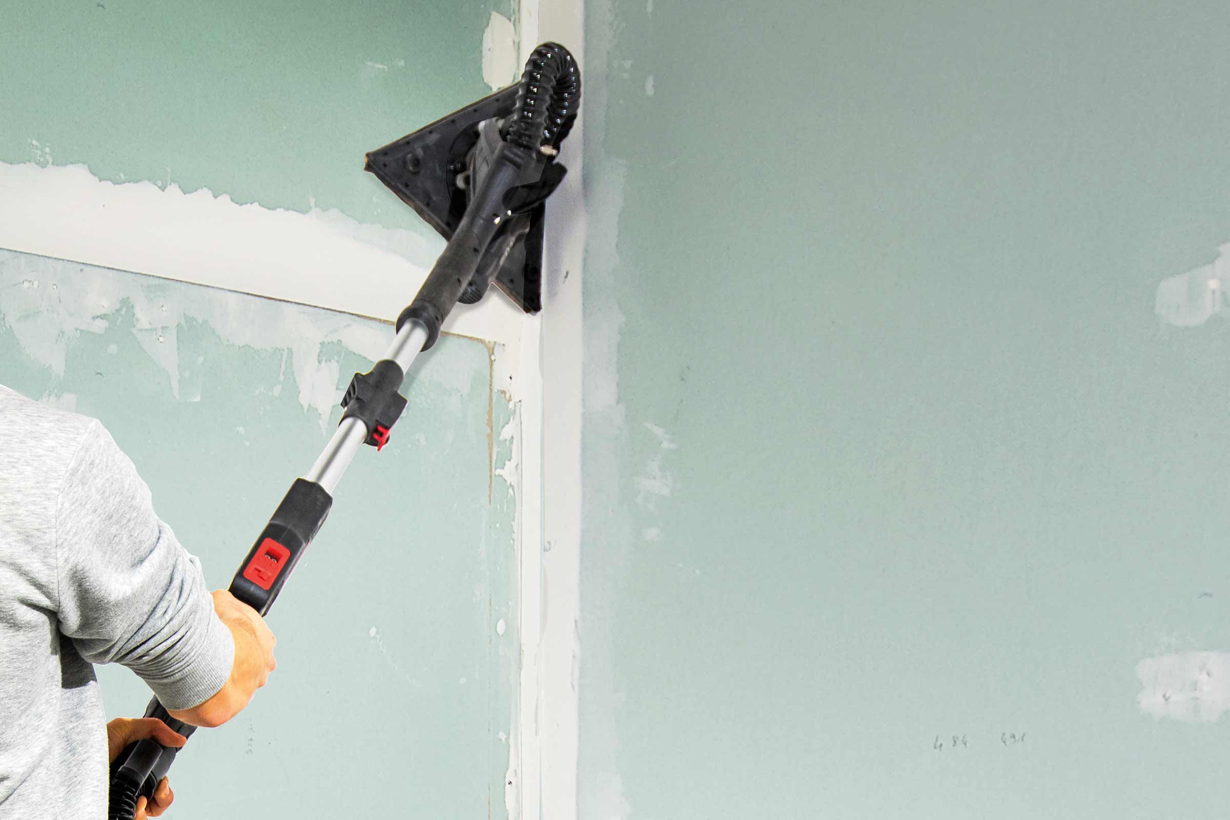 Een persoon schuurt een gipsplaten muur met een schuurmachine.