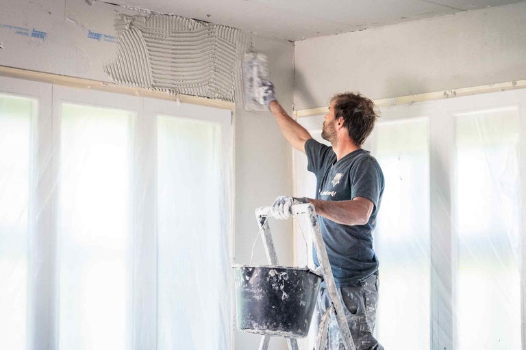 Een man op een ladder brengt met een troffel pleister aan op een muur boven een raam. Binnenopname.