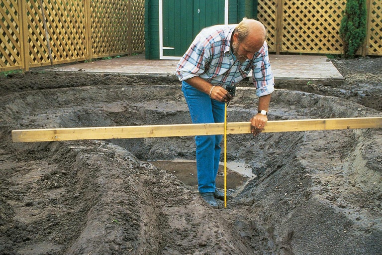Een man meet de diepte van een uitgraving voor een tuinvijver met een meetlint