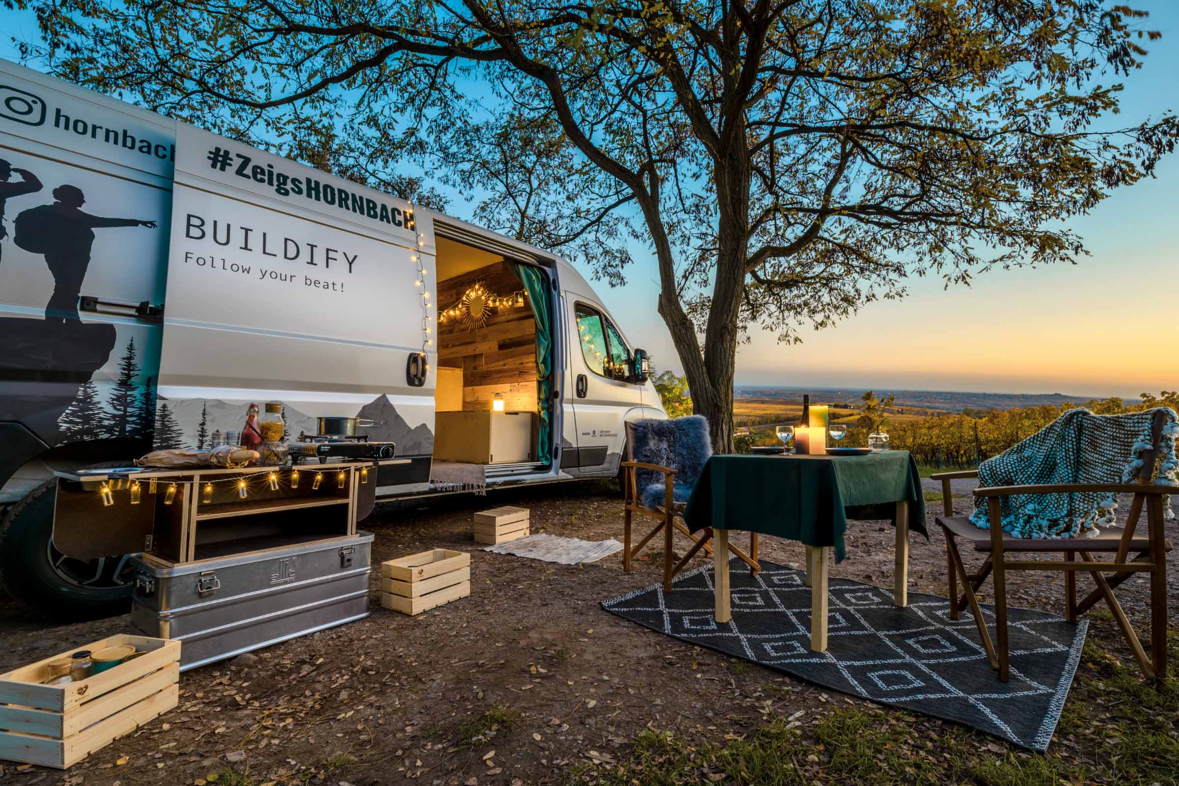Scene met een omgebouwde bus, een campingtafel en campingstoelen in de buitenlucht.