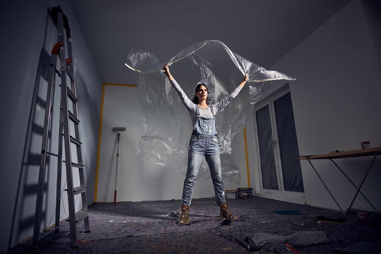 Een vrouw spant een beschermfolie in een kamer met afdekdoek, ladder en schildersbenodigdheden.