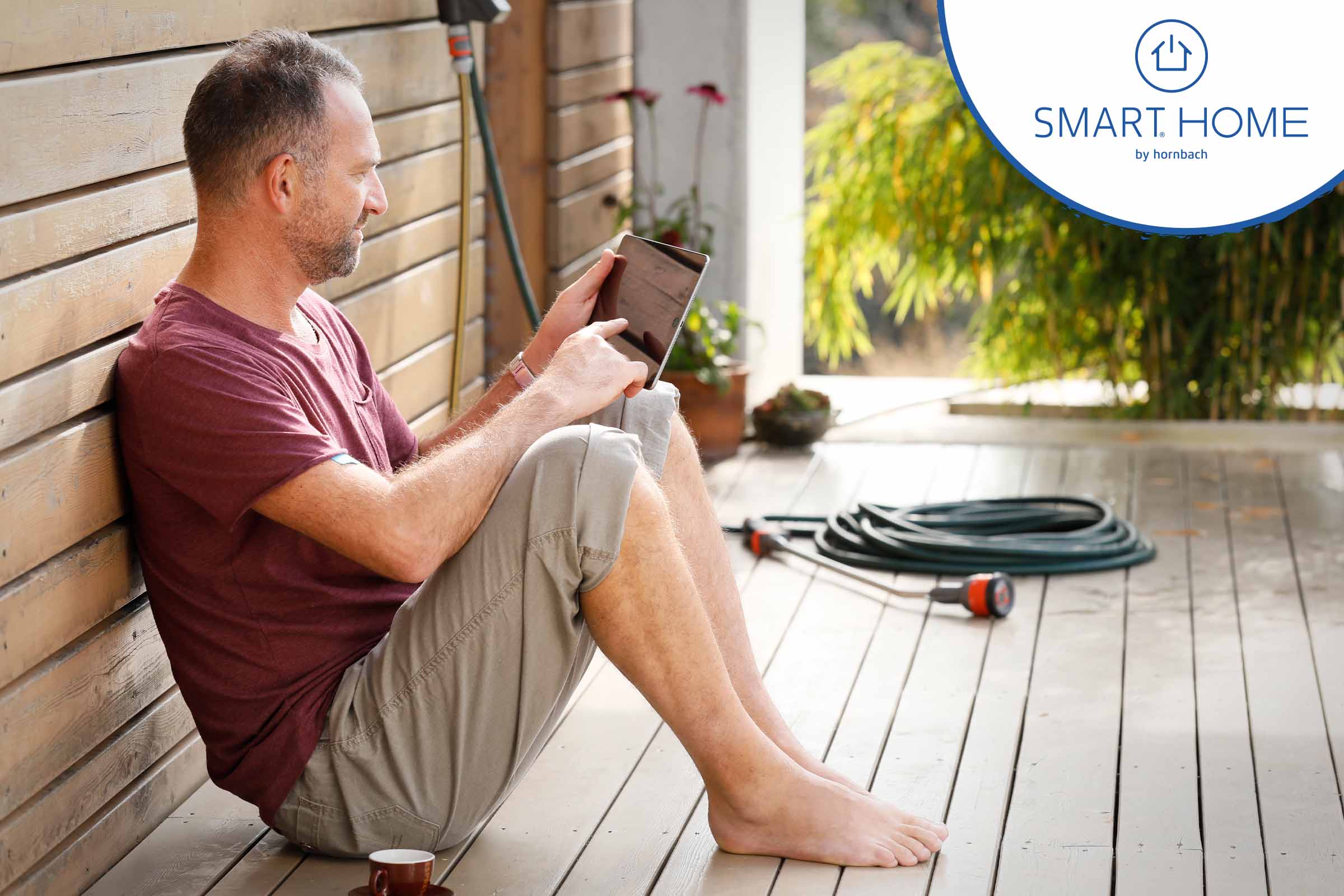 Man zit met tablet op een houten terras; tuinslang op de achtergrond; Hornbach Smart Home Logo