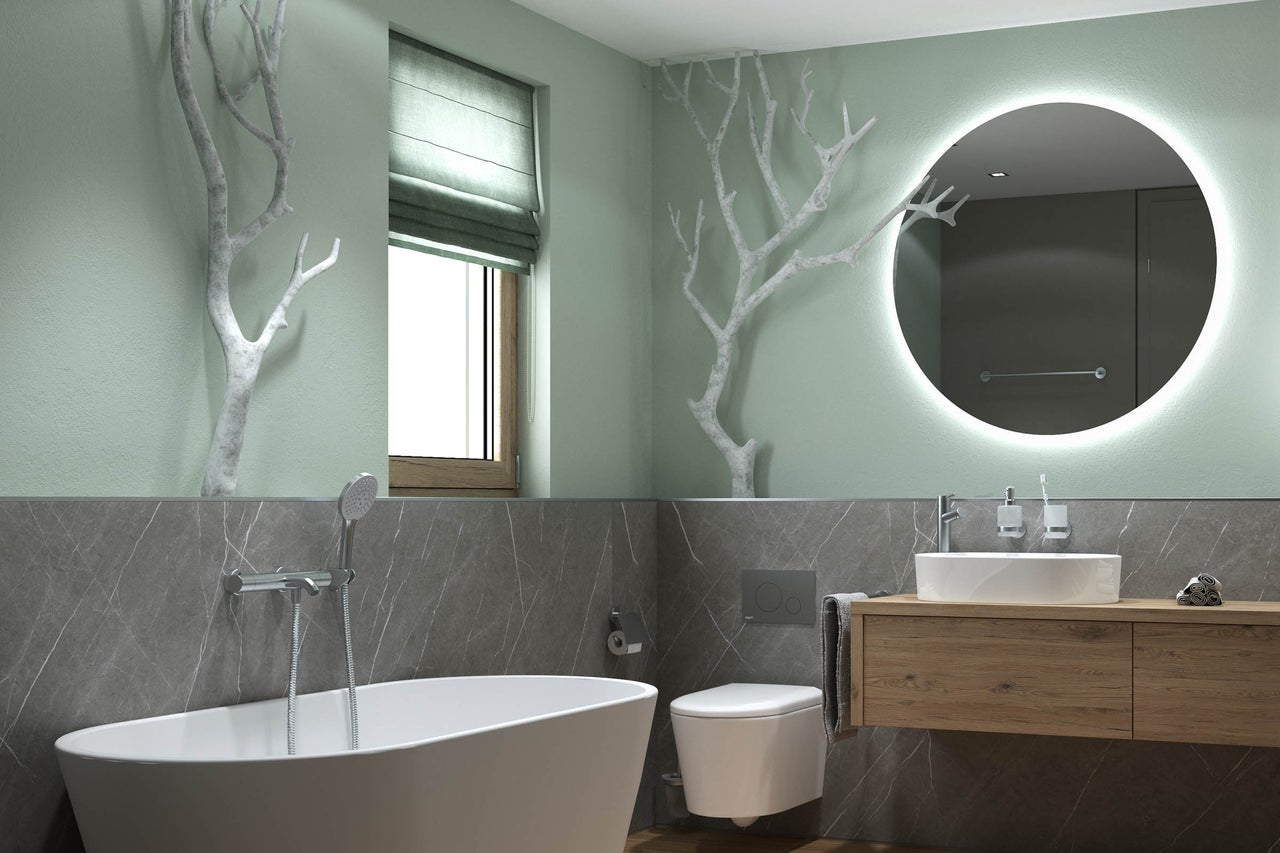 Moderne badkamer met vrijstaand bad, hangend toilet en houten wastafelmeubel. Grote ronde spiegel met verlichting en decoratieve takken aan de wand.