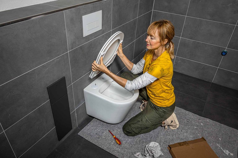 Een vrouw installeert een toiletbril in een badkamer met grijze tegels en een wandcloset.