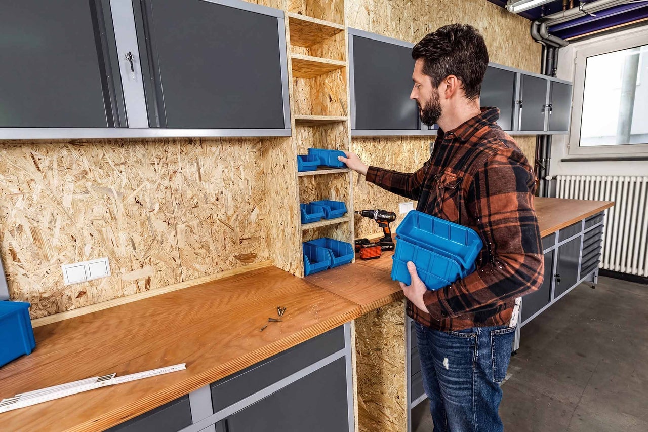 Man organiseert een werkplaats met blauwe magazijnbakken in een OSB-stelling boven een houten werkbank met grijze kasten.