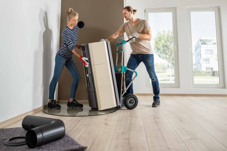 Twee mensen verplaatsen een houtkachel met een steekwagen in een kamer.