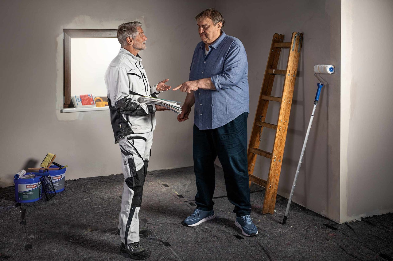 Twee mensen bespreken een renovatie met een ladder, verfroller en verfemmers in een kamer.