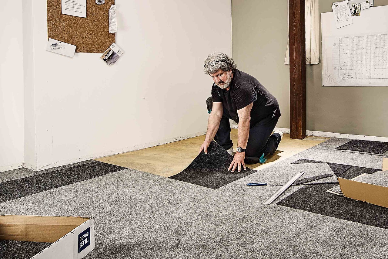 Een man legt tapijttegels in een kamer, waarbij hij grijze en donkere tapijttegels gebruikt