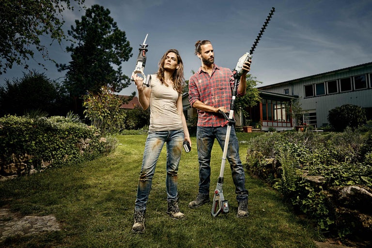 Een man en een vrouw staan met een heggenschaar en een reciprozaag in een tuin.