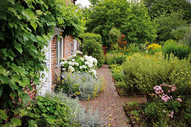 Verzorgde tuin met bloemen, struiken en bakstenen pad naast een bakstenen huis