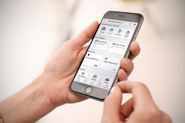 Een persoon bedient een smart home app op een smartphone voor het regelen van verlichting, zonwering en verschillende scenario's.