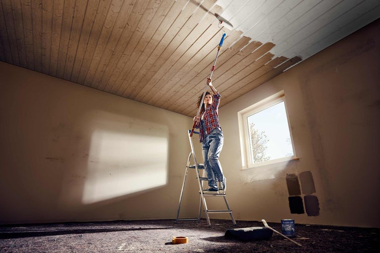 Vrouw schildert met een verfroller en telescopische steel een houten plafond vanaf een ladder. Verfbak, kwast en verfblik staan op de vloer.