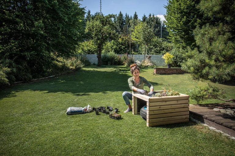 Een vrouw beplant een verhoogde tuinbak in de tuin, omgeven door tuingereedschap en gazon.