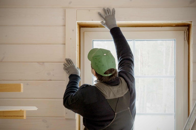 Ambachtsman monteert houten frame rond een raam in een kamer met houten lambrisering.