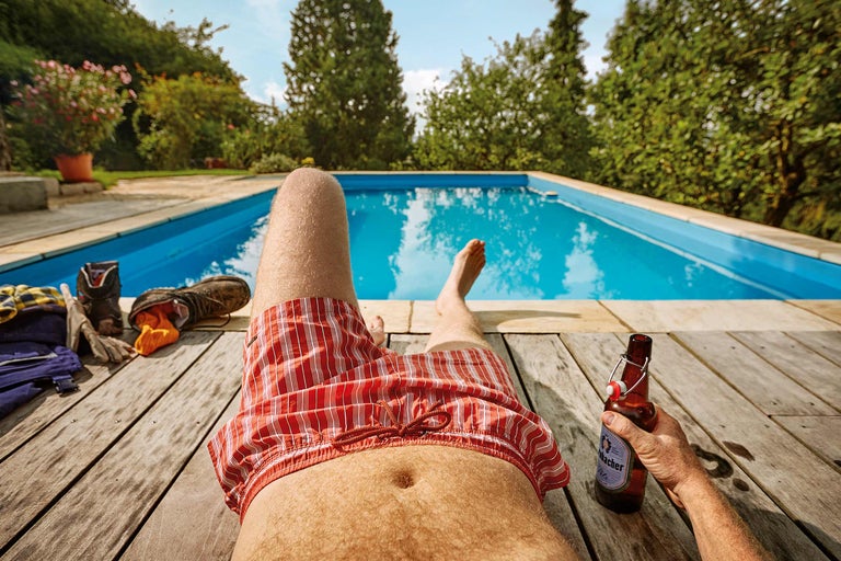 Man ontspant bij het zwembad met bier, werkbroek en schoenen op houten terras.