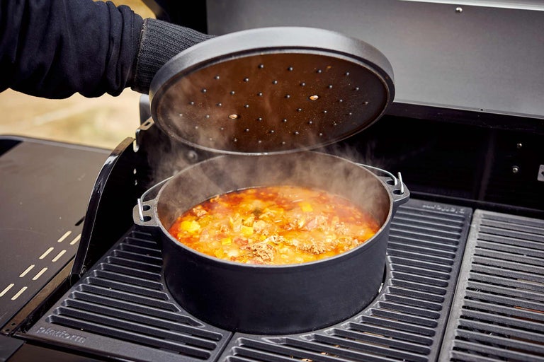 Een Dutch Oven met deksel staat op een grill, gevuld met een gerecht waar stoom vanaf komt.