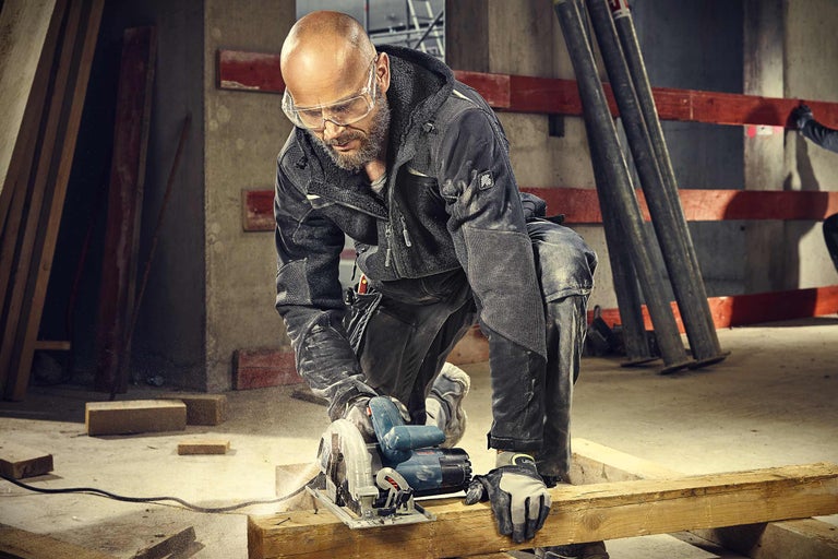 Een vakman zaagt een houten plank met een handzaagmachine. De man draagt werkkleding en een veiligheidsbril.