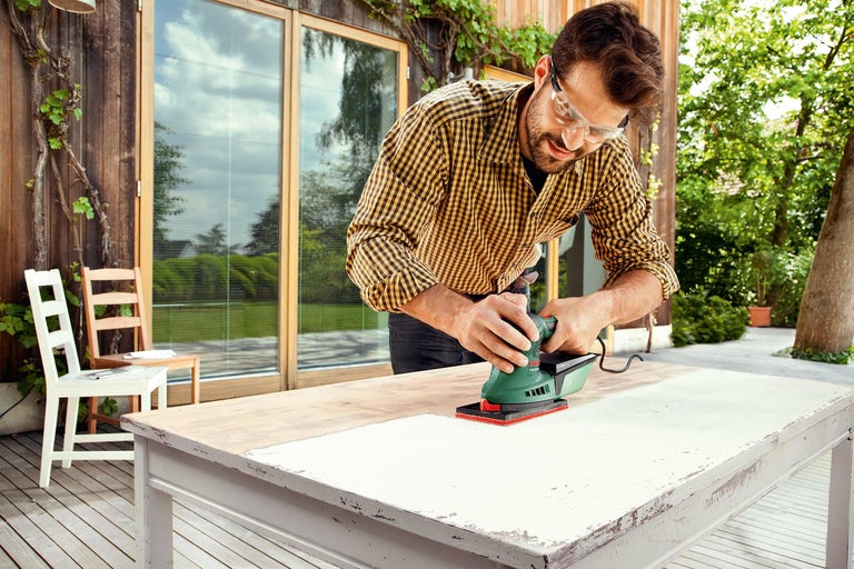 Een man schuurt een tafelblad met een schuurmachine in de buitenlucht.