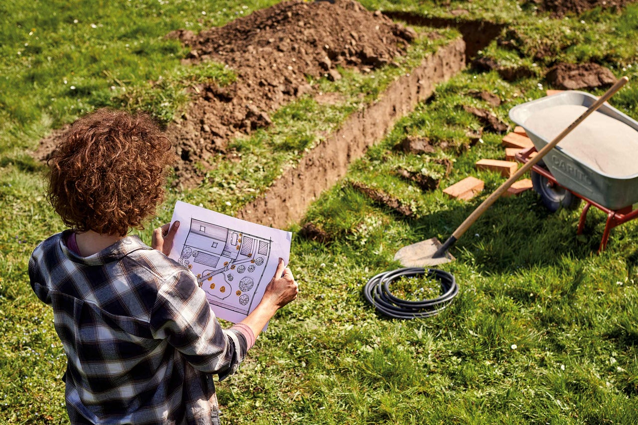 Vrouw bekijkt bouwplan in tuin met schop, kruiwagen, slang en uitgegraven greppel