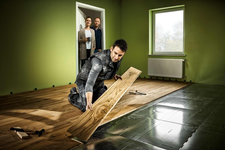 Een man legt laminaatvloer in een kamer met een raam en een radiator. Op de achtergrond staan twee mensen in een open deur.