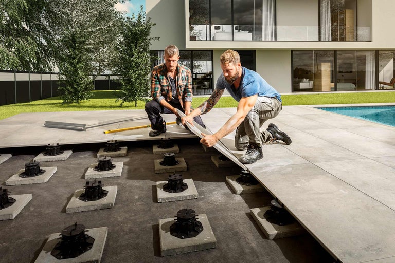 Twee vakmannen leggen terrastegels op tegeldragers in een tuin.