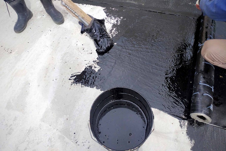 Bitumen coating wordt aangebracht met een borstel, dakbedekkingsbaan en emmer met bitumen coating zijn ook zichtbaar