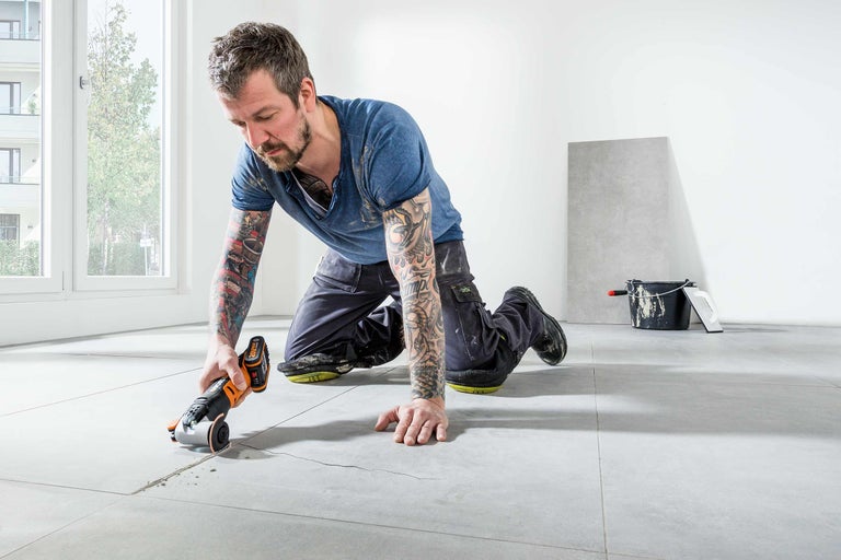 Man knielt op tegels en werkt met een multifunctioneel gereedschap aan een beschadigde voeg.