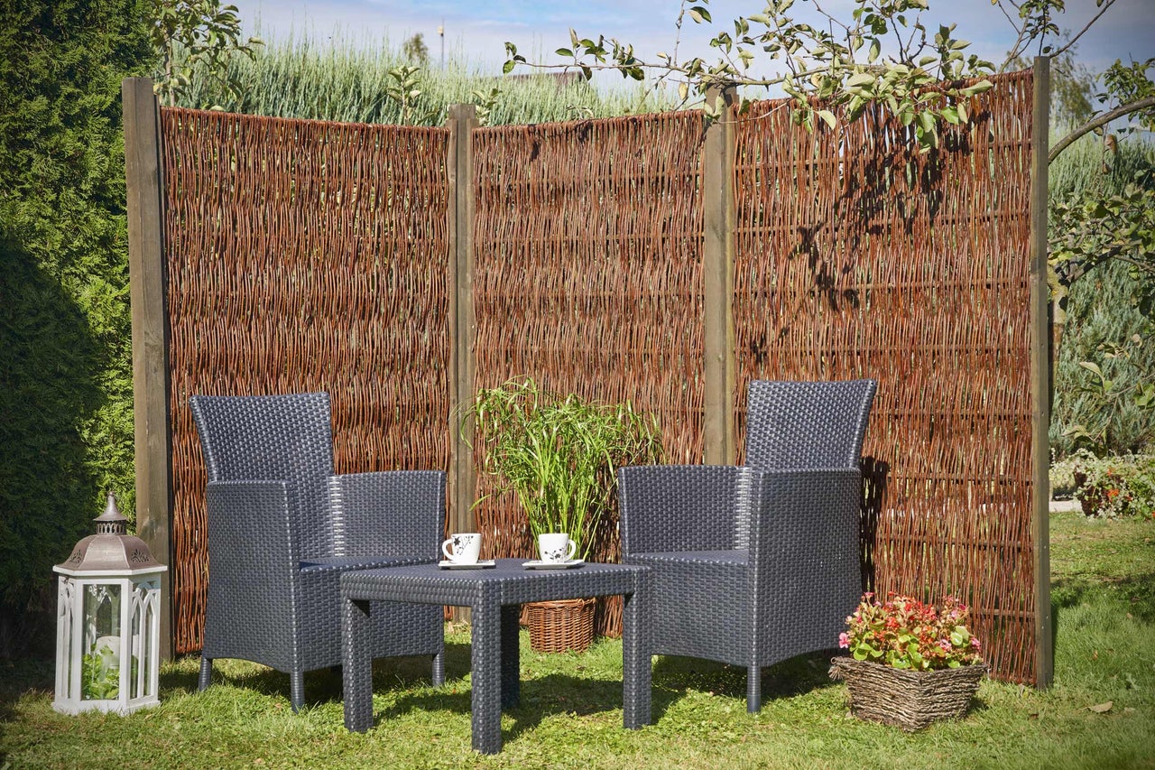 Tuinscène met wilgentenen schutting, rotan stoelen en tafel op een gazon.