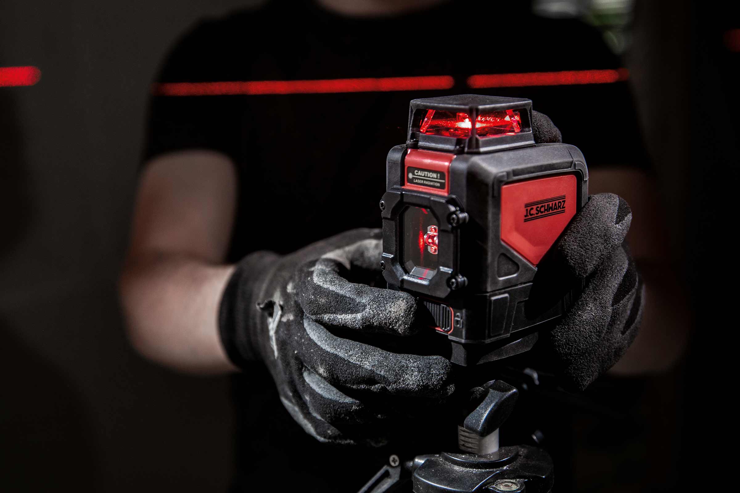Persoon met werkhandschoenen houdt een J.C. Schwarz kruislijnlaser op een statief vast, die rode laserlijnen projecteert.