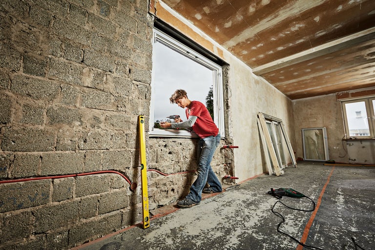 Een man monteert met een accuschroevendraaier een raam in een casco woning. Een waterpas leunt tegen de muur.