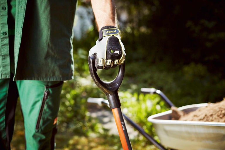 Een persoon met werkhandschoenen houdt een spade vast met een gevulde kruiwagen op de achtergrond.