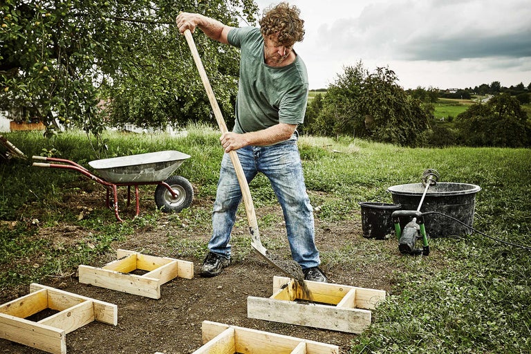 Een man vult houten frames met een schop in de tuin, terwijl een kruiwagen en een speciekuip klaarstaan.