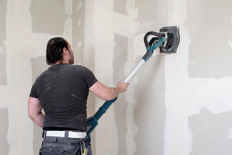 Ambachtsman schuurt een muur met een schuurmachine met lange steel. De ruimte ziet er licht en stoffig uit.