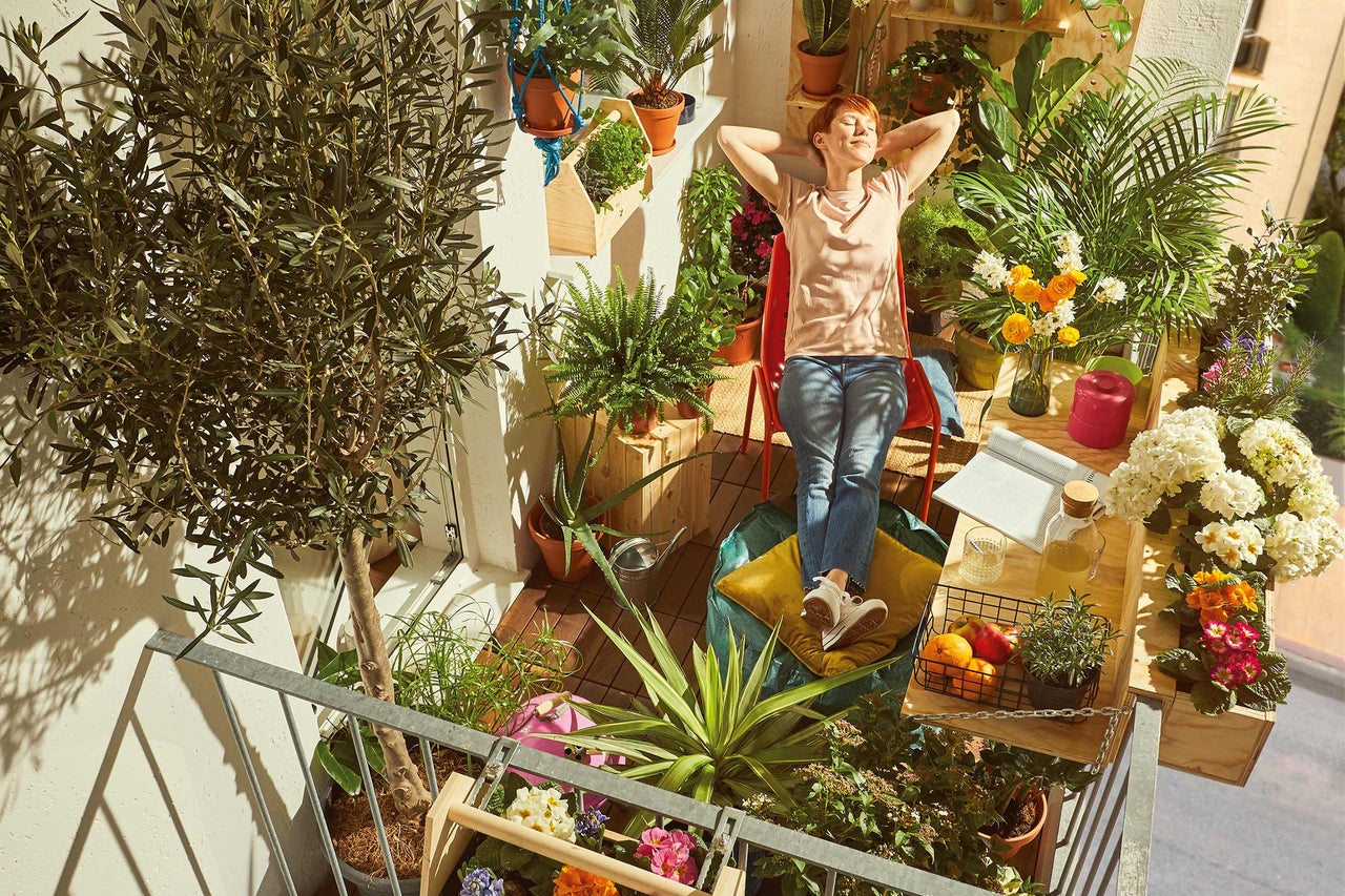 Een vrouw ontspant op een groen balkon, omringd door planten in potten en bakken, met een tafel met drankjes en fruit.
