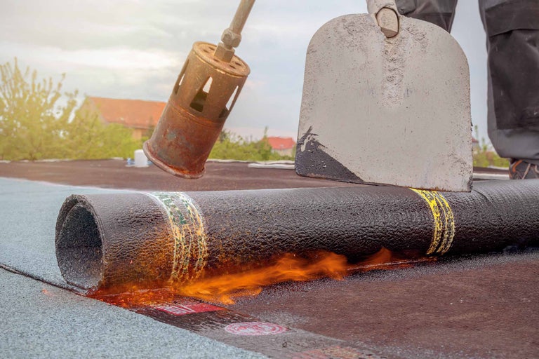 Dakdekker werkt met een dakbrander en een schep aan een bitumen dakbedekking op een dak.