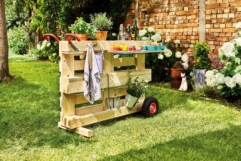 Een mobiele houten tuinbar met wielen en een werkblad staat in de tuin.