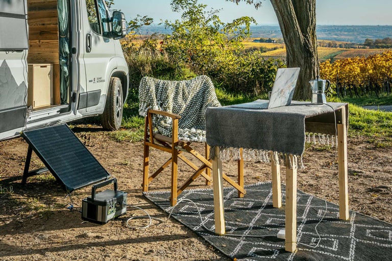 Buitenscène met camper, zonnepaneel, tafel, stoel en laptop in een natuurlijke omgeving.