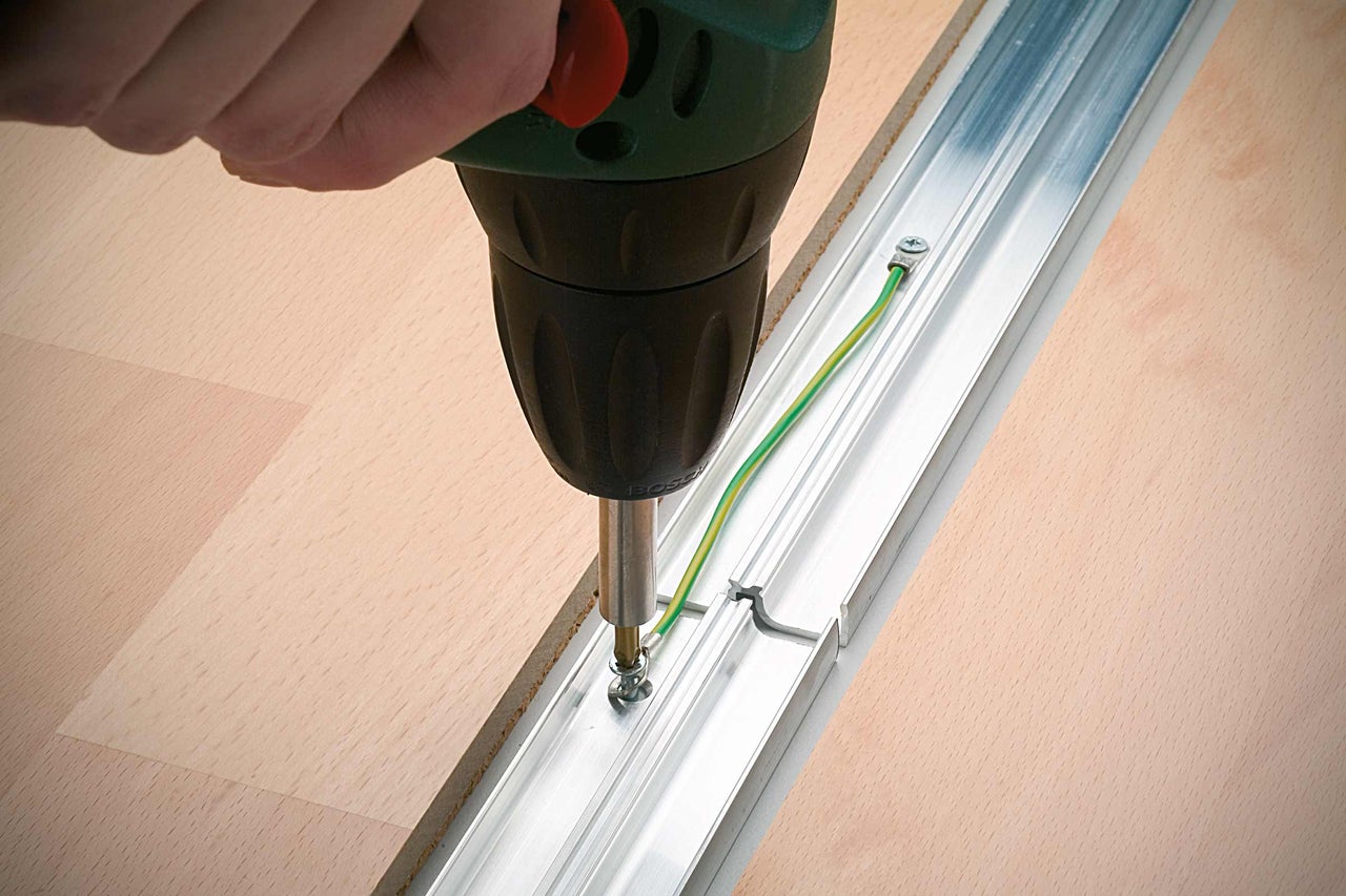 Een persoon schroeft met een accuboormachine een aluminium profielrail op een houten vloer.