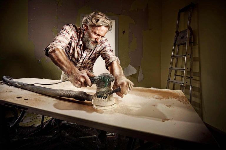 Een man schuurt een houten blad met een schuurmachine in een kamer met een ladder.