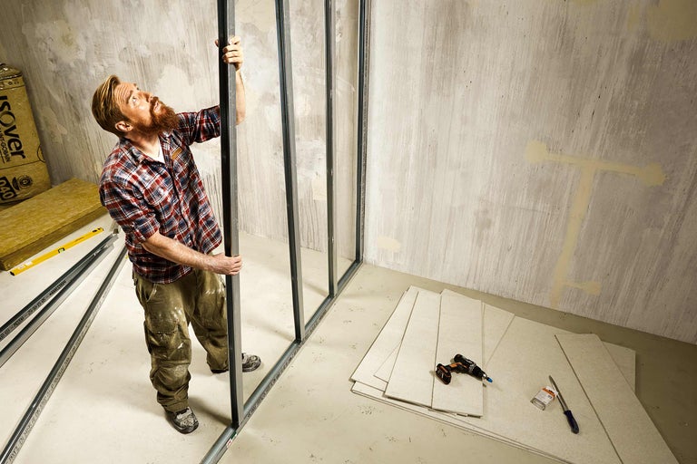 Een man monteert metalen profielen voor een gipsplatenwand in een ruimte met gipsplaten, isolatie en gereedschap.