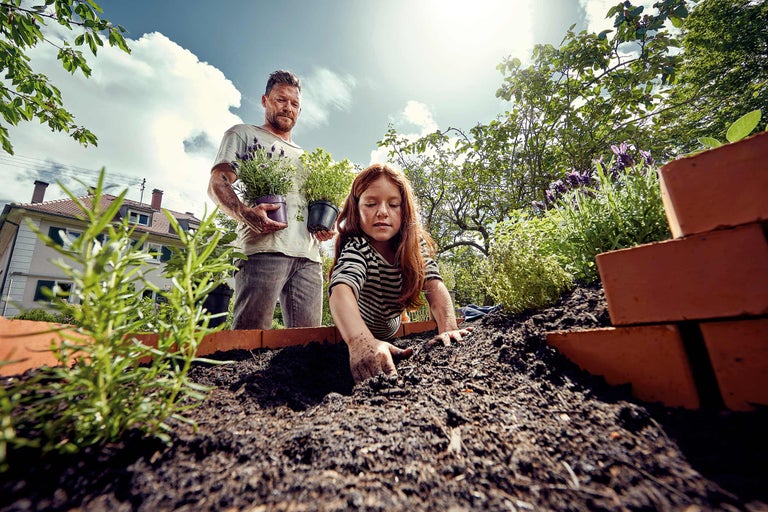 Vader en dochter beplanten een verhoogde tuinbak met kruiden en aarde in een tuin.