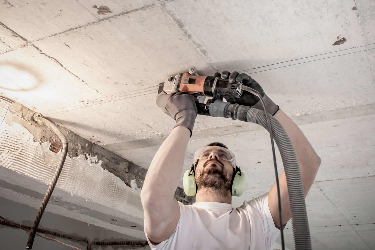 Een man schuurt een betonnen plafond met een betonmolenmachine, terwijl hij een veiligheidsbril en gehoorbescherming draagt