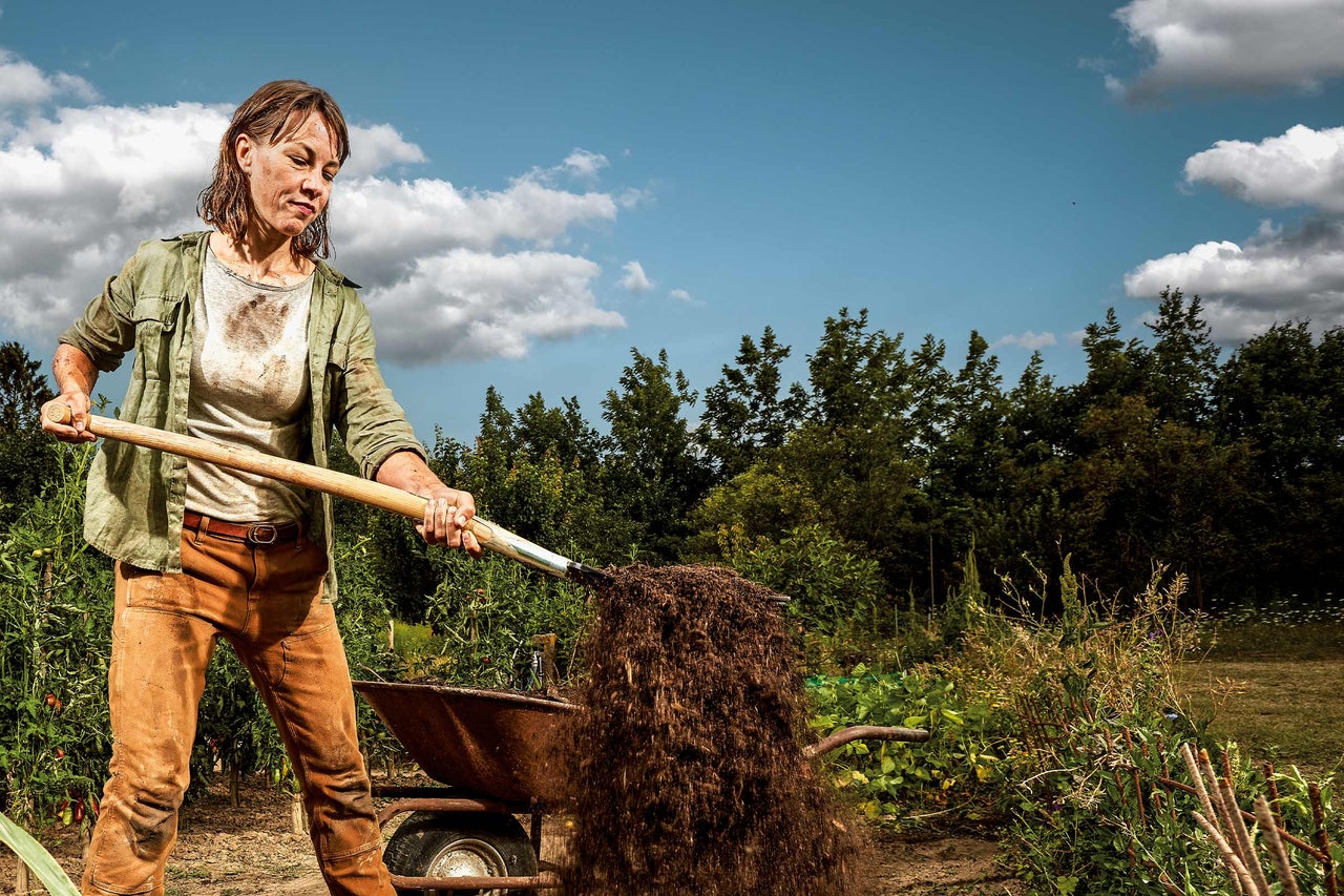 Een vrouw schept aarde met een schop in een kruiwagen in een tuin.