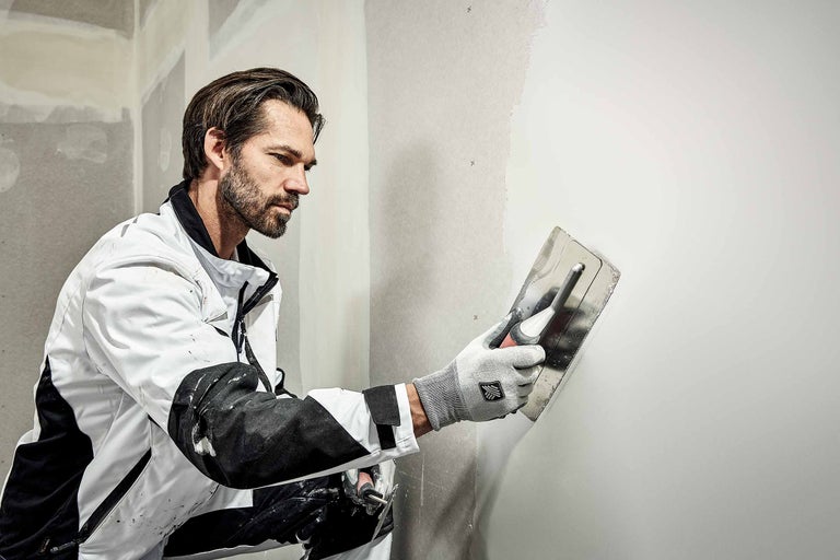 Een man egaliseert een muur met een pleisterspaan. De muur is gedeeltelijk gepleisterd.