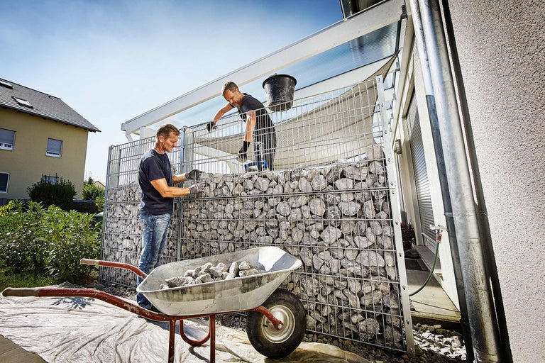 Twee mannen bouwen een schanskorfmuur met stenen, een kruiwagen is gevuld met stenen.