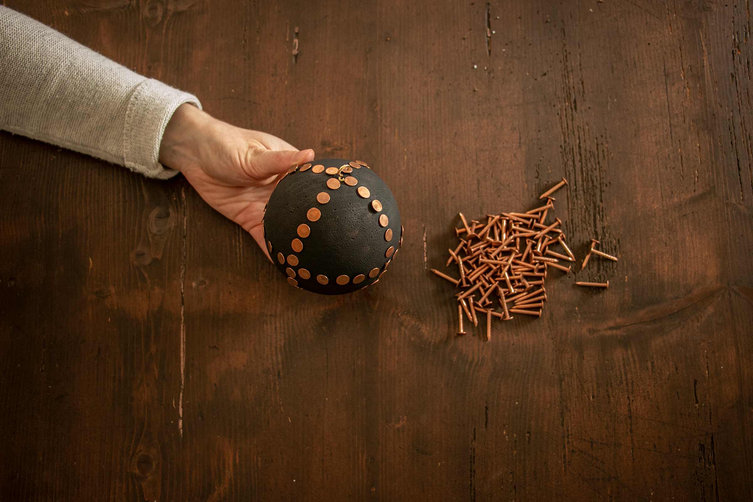 Een hand houdt een zwarte bol met koperen klinknagels vast op een houten tafel naast losse koperen klinknagels.