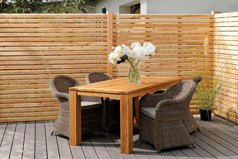 Scène met houten tafel, rotan stoelen en houten schutting op een terras.